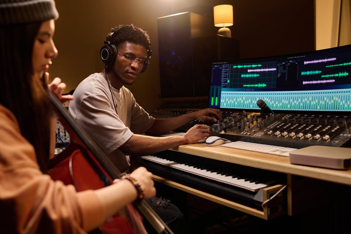 Young Black man operating audio mixing console while young Latin woman plays cello in professional recording studio