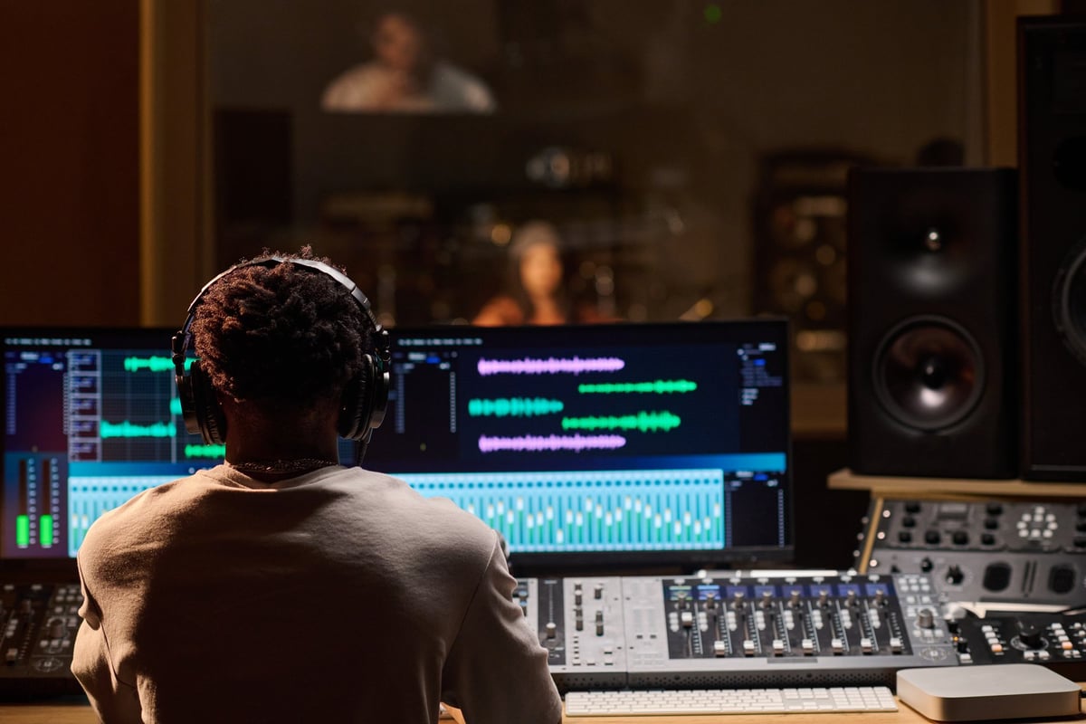 Black man wearing headphones operating audio mixing console and computer monitors in professional recording studio