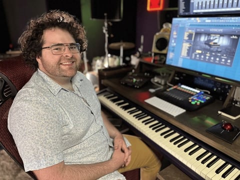 Man smiling at camera in music production studio with keyboard and monitor displaying audio software
