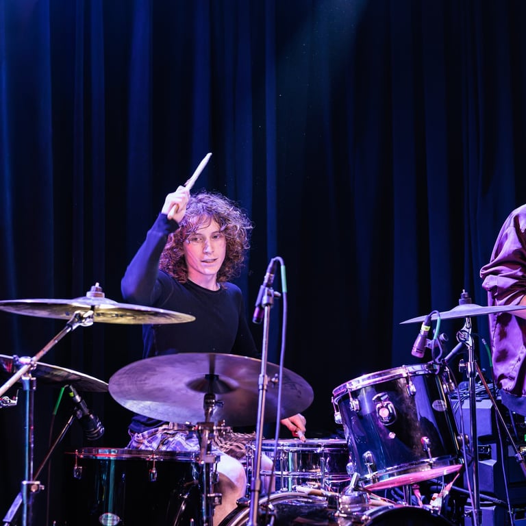 A curly-haired drummer performs on stage with drumsticks raised, illuminated by purple stage lighting against a dark blue curtain backdrop.