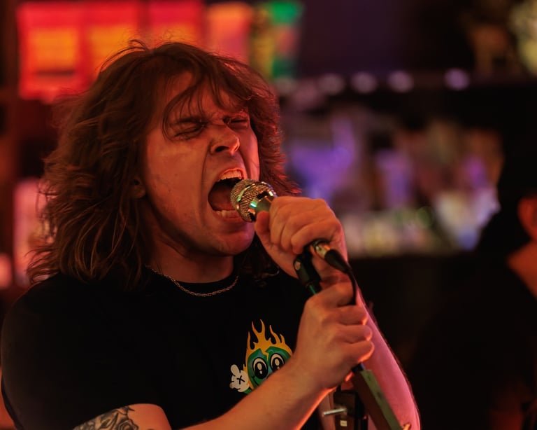 Musician with long dark hair passionately singing into microphone on stage with colorful red and purple stage lighting