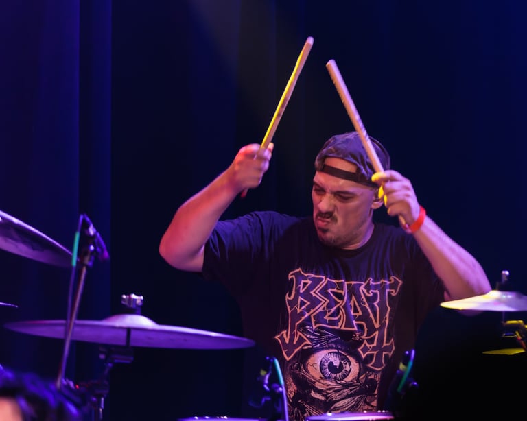 Drummer performing on stage with drumsticks raised, wearing black band t-shirt and headband, purple stage lighting
