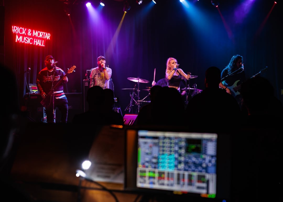 Live band performing on stage with colorful purple and blue lighting, audience in foreground with audio equipment visible