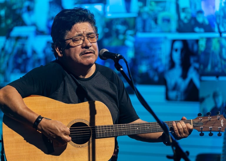 Man wearing glasses playing acoustic guitar at microphone on stage with blue lighting in background