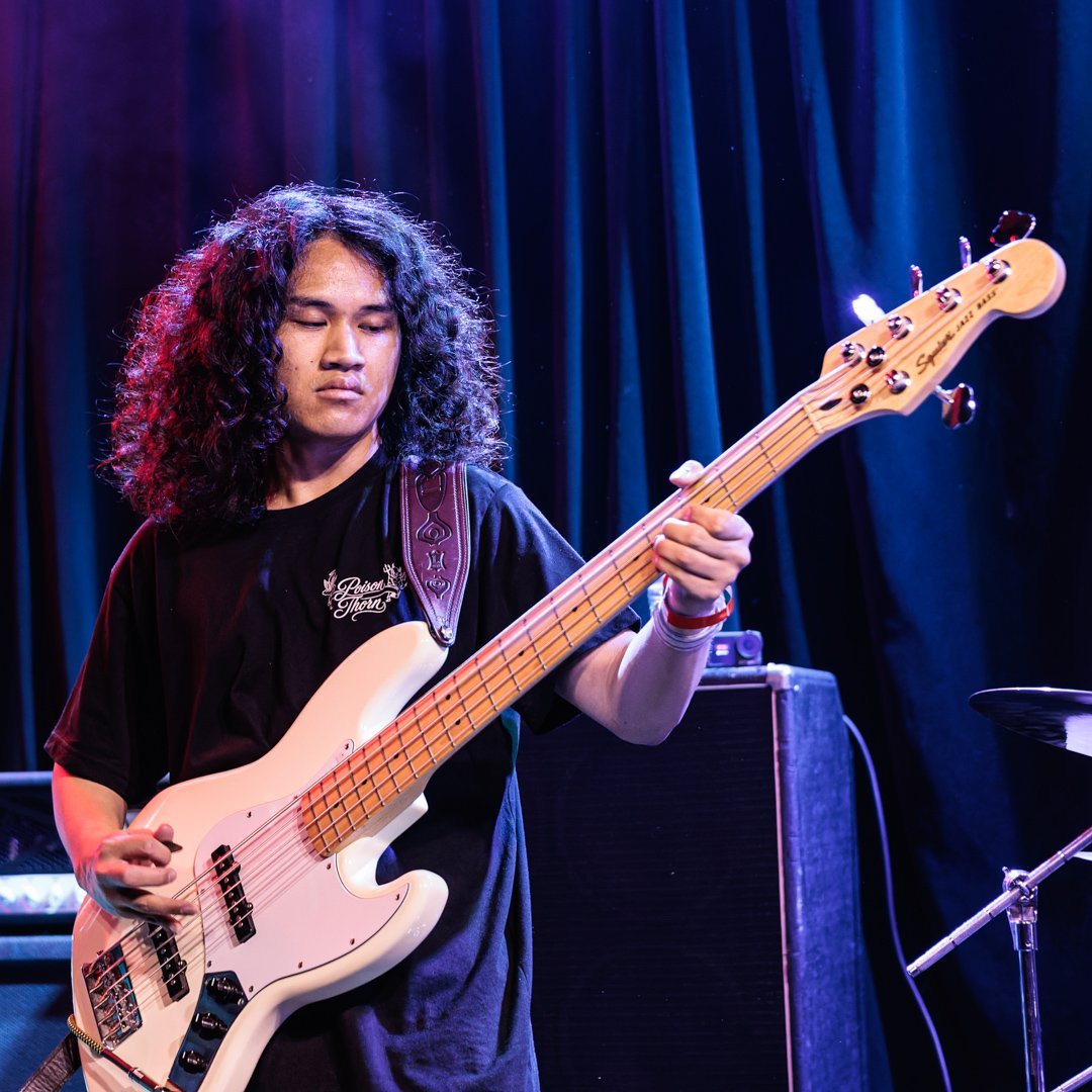 Musician with long curly hair playing electric bass guitar on stage with blue curtain background