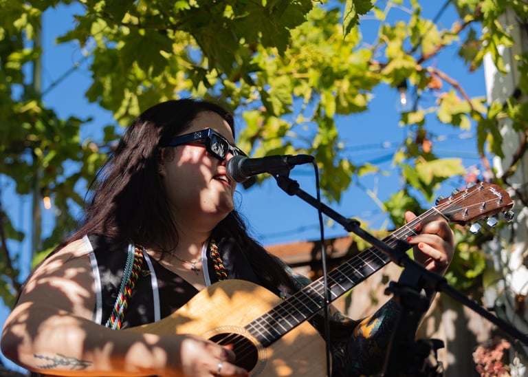 Male musician performing outdoors with acoustic guitar under green foliage and blue sky