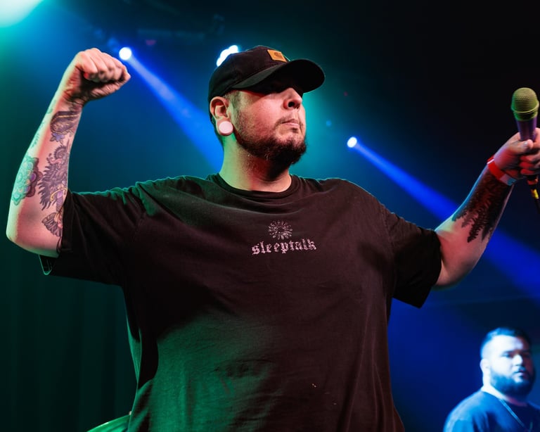 Man performing on stage wearing black t-shirt and cap, with fist raised and microphone in hand against blue stage lighting
