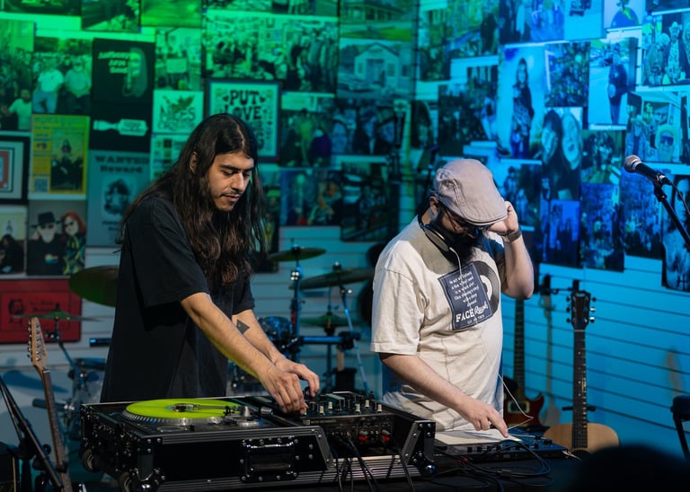Two DJs performing at turntables with neon-lit wall of photographs in the background