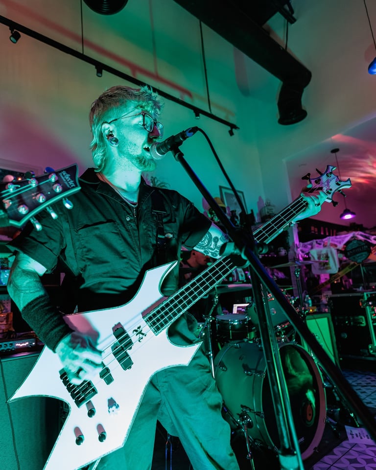 Musician performing on white bass guitar under turquoise and pink stage lighting with microphone and band equipment visible