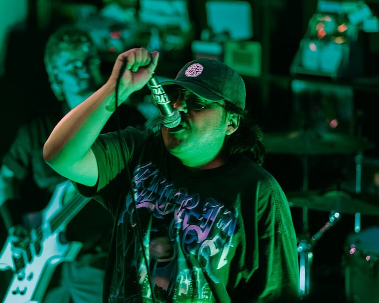 Person performing on stage wearing a black cap and graphic t-shirt, adjusting their glasses under green stage lighting