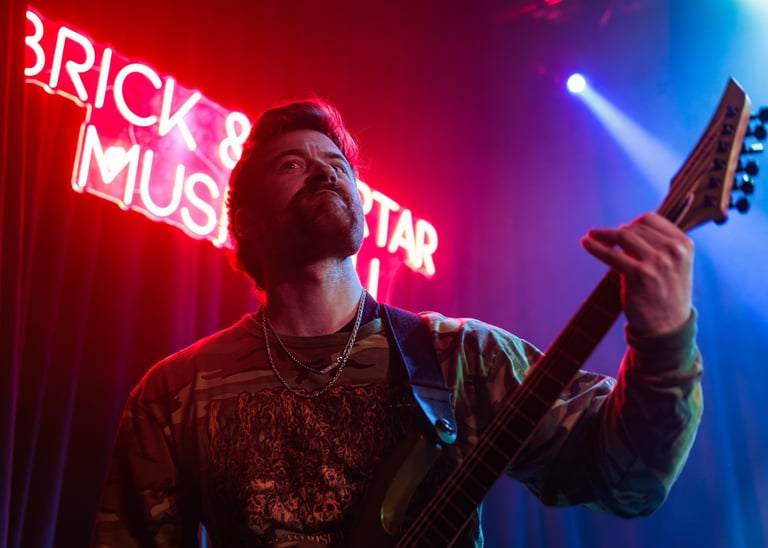 Bearded musician playing electric guitar on stage with red and blue neon lights and Brick Mustard sign in background