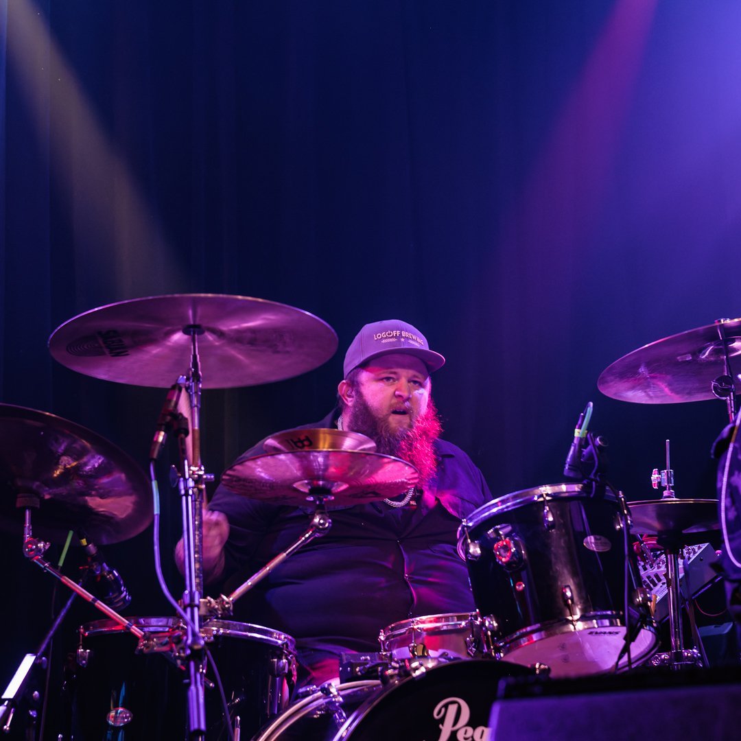 Drummer performing on stage with purple lighting, wearing a cap and playing a full drum kit with cymbals.