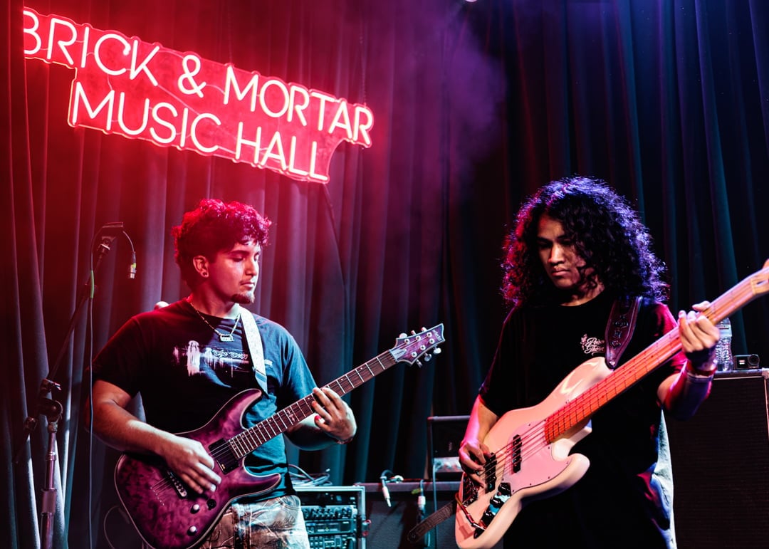 Two musicians perform on stage at Brick and Mortar Music Hall with red neon signage, one playing electric guitar on the left and another on bass guitar on the right
