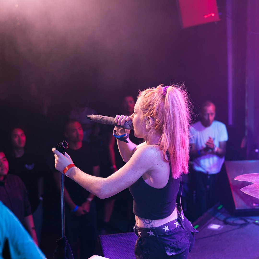 Female performer singing into microphone on stage under purple lighting with audience members visible in background
