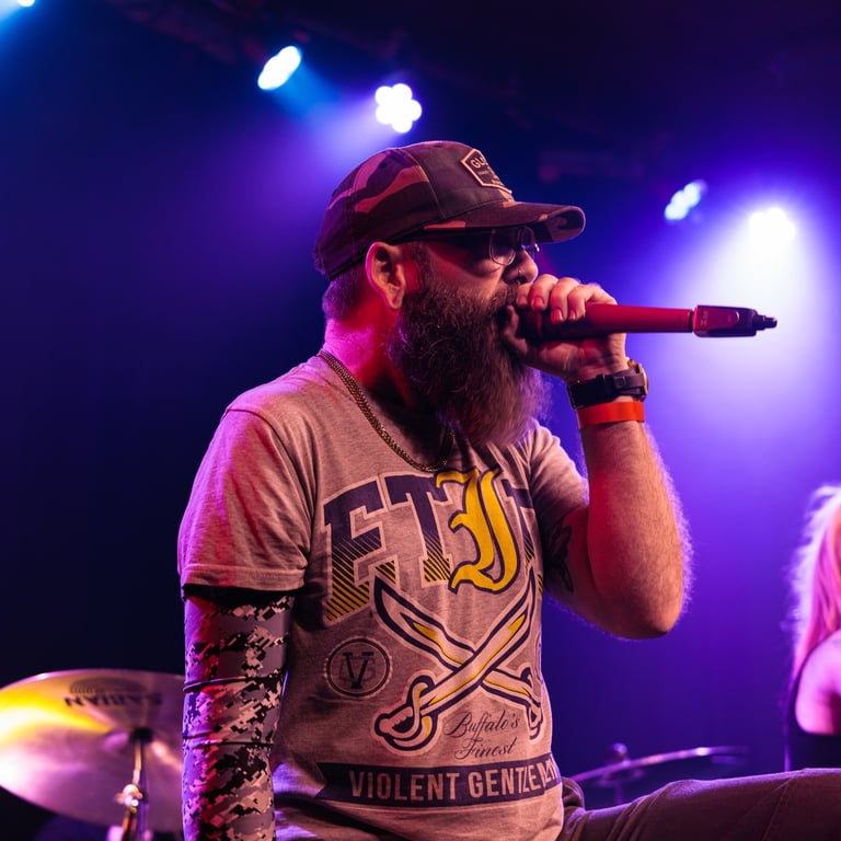 Bearded male performer singing into microphone on stage under purple and blue stage lights, wearing baseball cap and graphic t-shirt