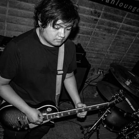 Black and white photo of a young man playing electric guitar on stage, wearing dark clothing with a lanyard