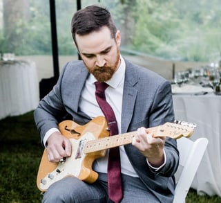 Bearded man in business suit and burgundy tie playing acoustic electric guitar outdoors