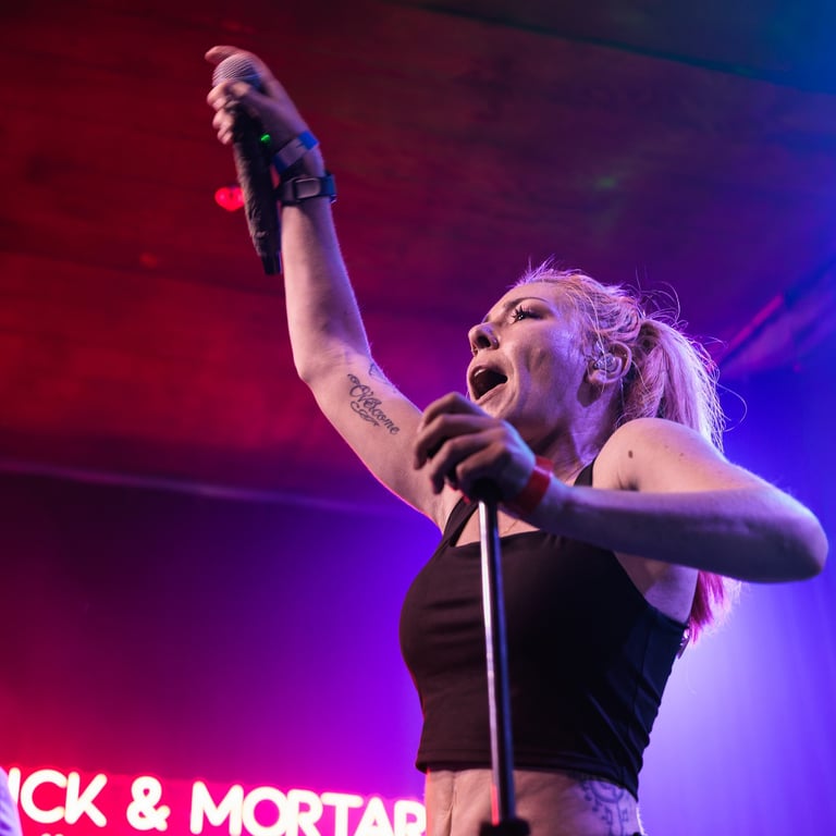 Female singer performing on stage with fist raised, holding microphone, wearing black top under red and blue stage lighting