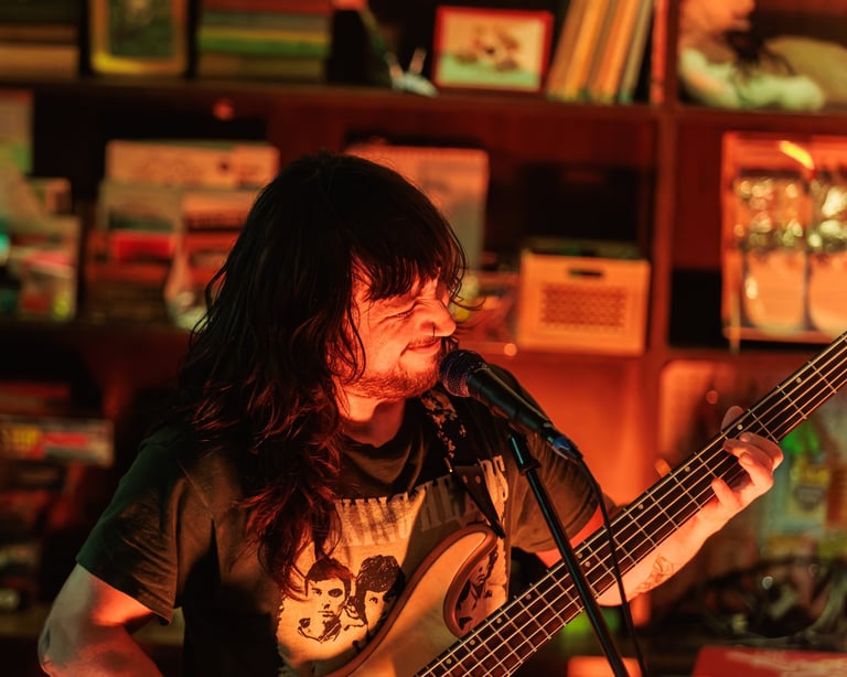Musician with long dark hair playing bass guitar and singing into microphone on stage with warm orange lighting