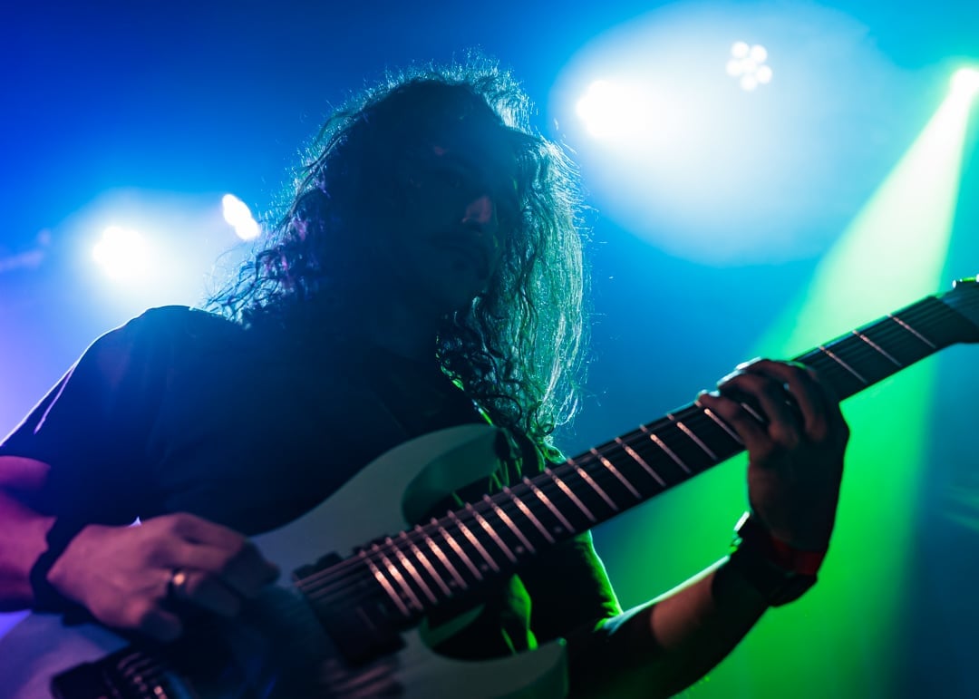 Musician with long hair playing electric guitar on stage under blue and green stage lights
