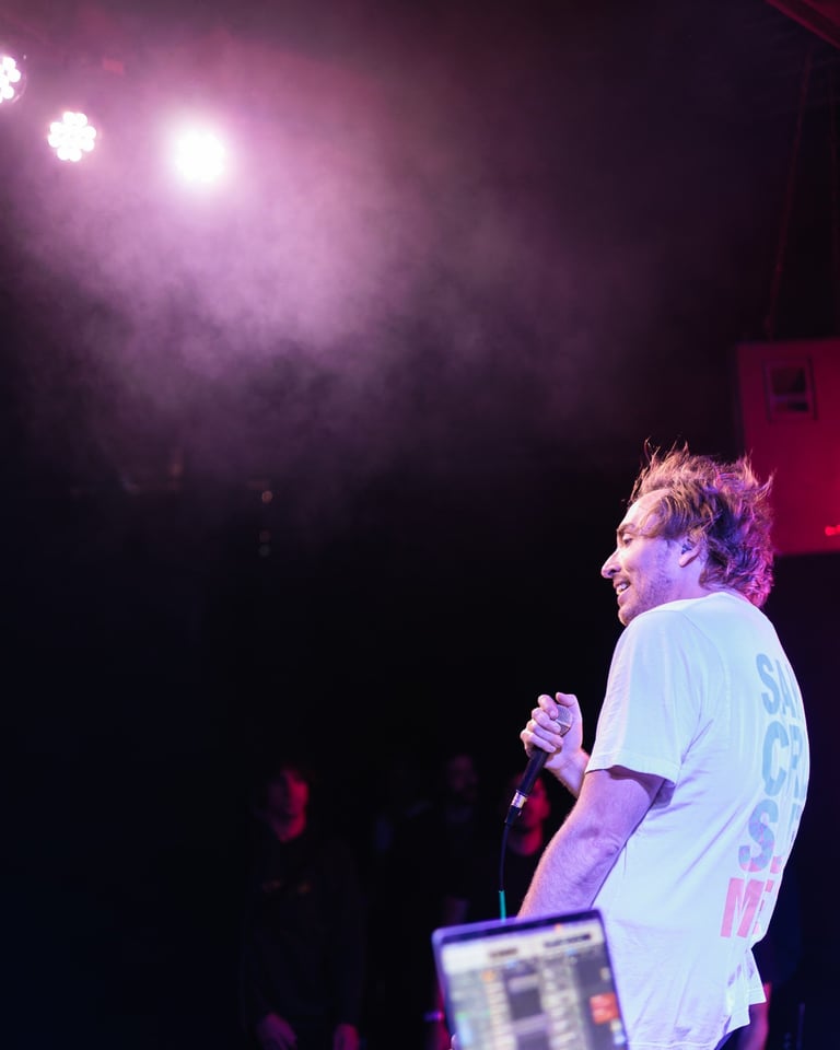 Man performing on stage with microphone, wearing light blue shirt, purple stage lighting in background