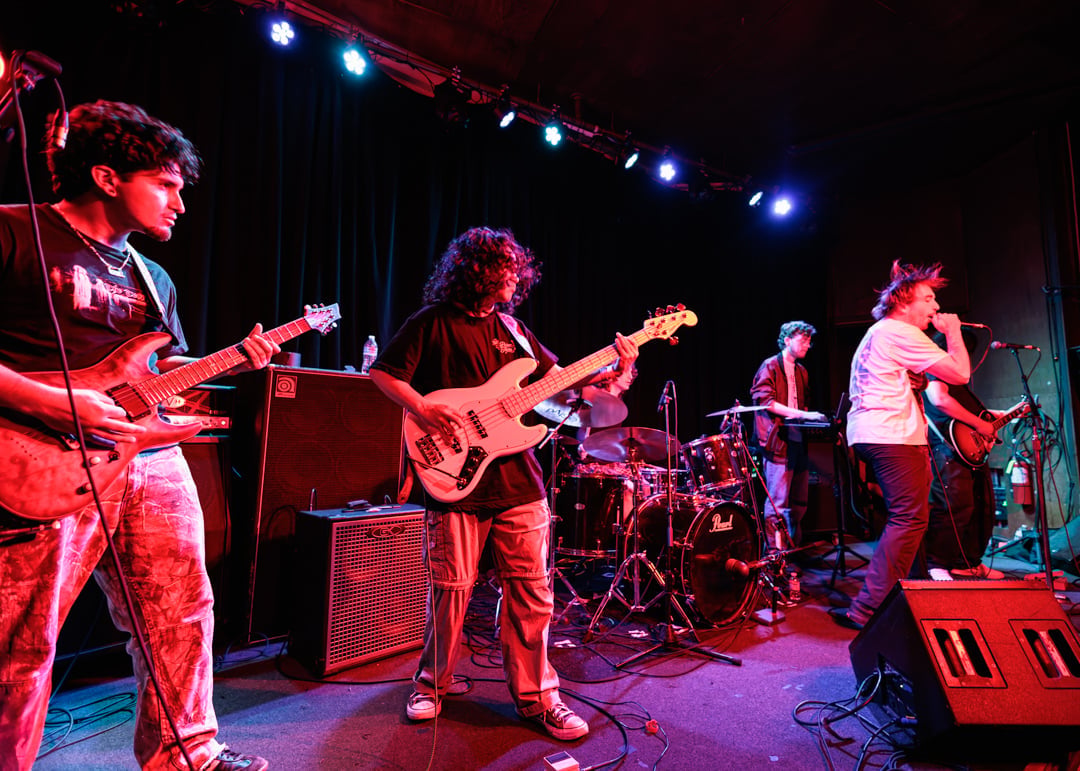 Live band performing on stage with guitarists, bassist, and vocalist under colorful red and blue stage lighting