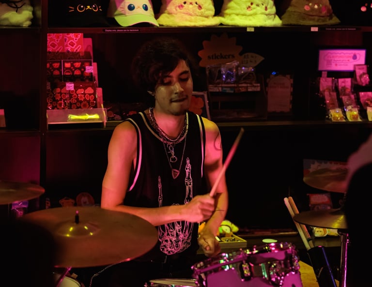 Musician playing drums in neon-lit room with plush toys and arcade items on shelves behind him