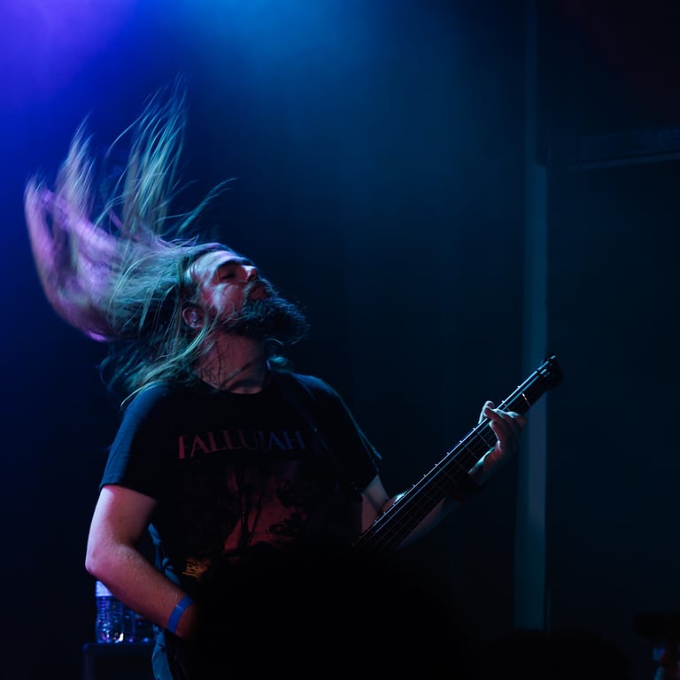 Musician with long blonde hair playing electric bass guitar on stage under blue and purple stage lighting