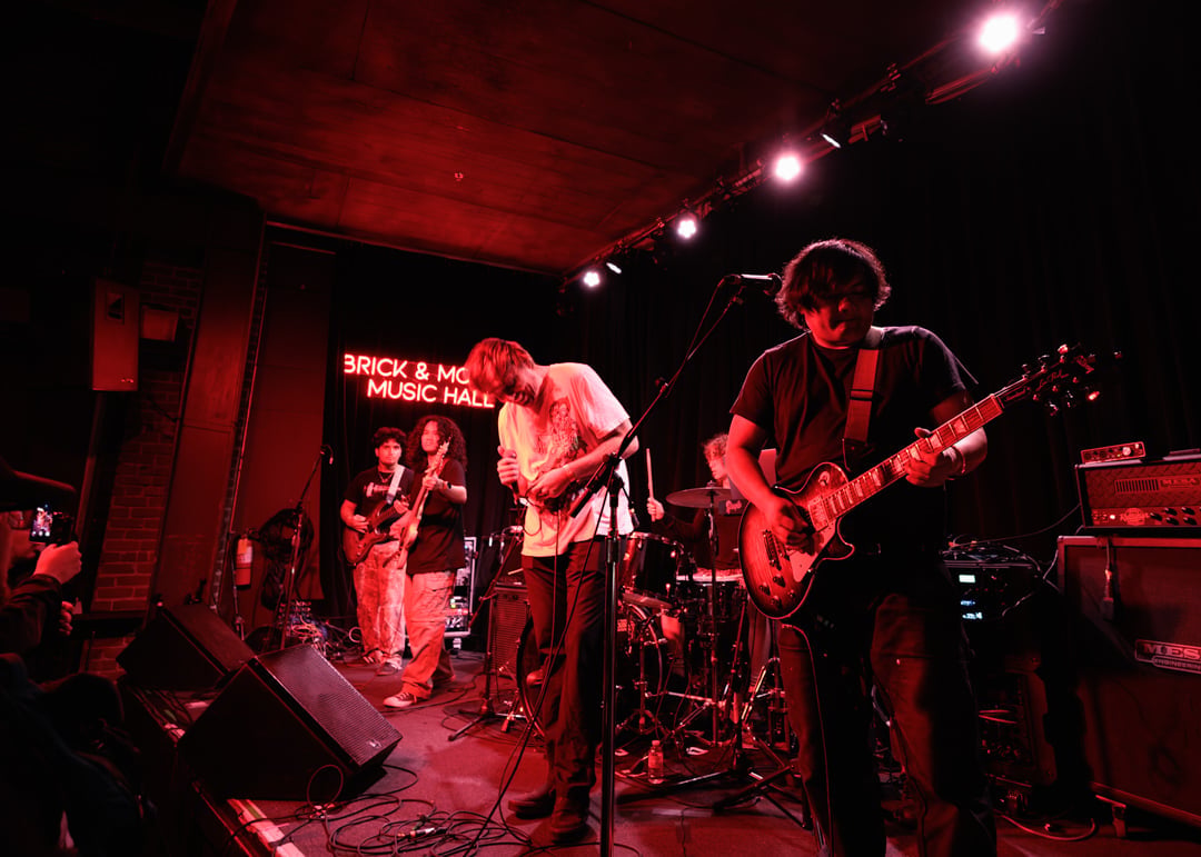 Live band performing on stage in a dimly lit venue with red lighting, featuring guitarists and drummer