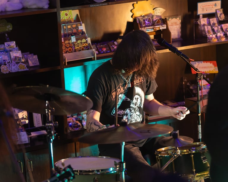 Drummer performing on stage wearing a black t-shirt in a dimly lit venue with neon lighting and band merchandise displayed on walls