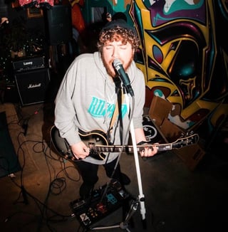 Musician with curly hair performing with electric guitar at microphone on stage with colorful graffiti backdrop